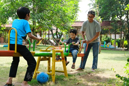 Preview: Joyful scene of a father and his two sons playing on a colorful seesaw in a lush, green park.