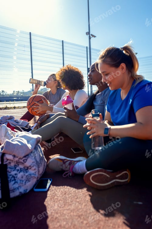 Preview: Diverse group of young woman sitting on court resting afrer playing basketball outdoors.