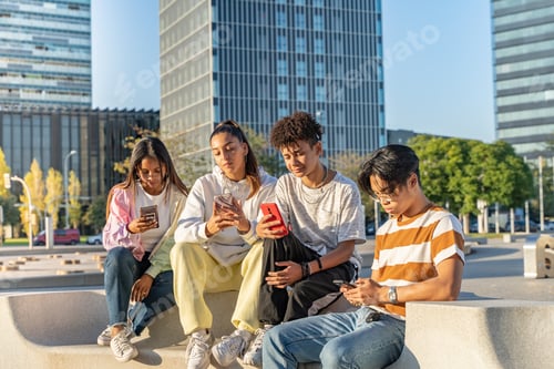 Preview: Group of happy teenage friends looking the phone and laughing in a bench in the city street.