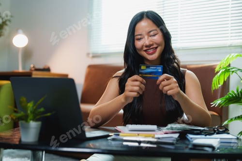 Preview: Smiling asian freelancer holding credit card and using laptop for online shopping at home
