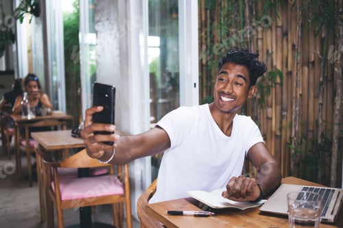 Preview: Cheerful ethnic man taking selfie while working on laptop in cafe