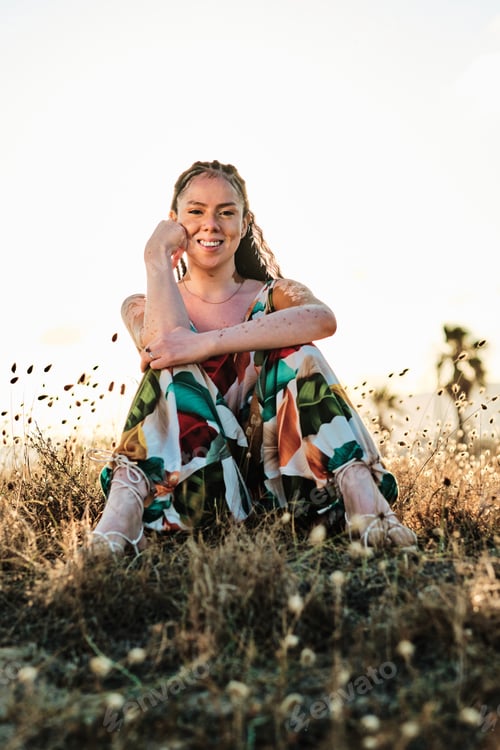 Preview: Woman with vitiligo looking at camera and smiling while sitting outdoors in nature.