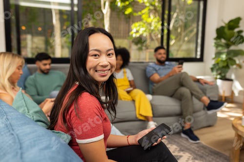 Preview: Smiling woman holding smartphone, sitting with friends on couch in living room