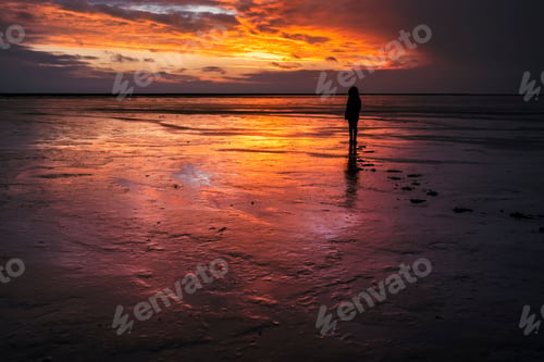 Preview: Silhouette of a person standing in shallow water against the sea at orange sunset