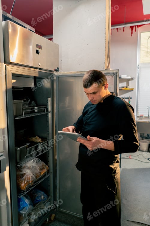 Preview: chef stands in kitchen in a black uniform and looks through what is on the list in the refrigerator