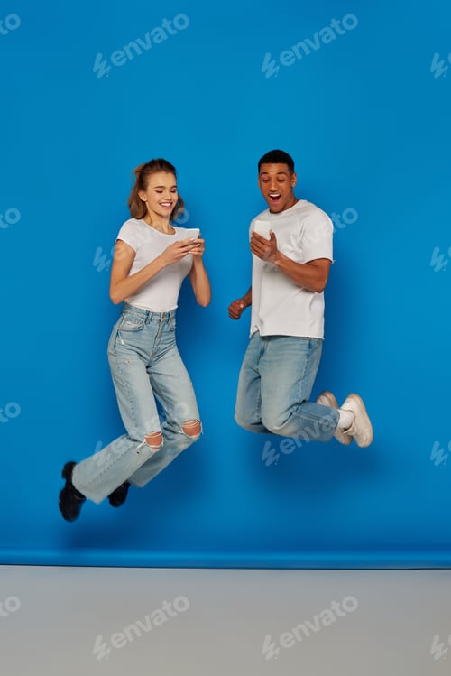 Preview: happy multicultural couple using smartphones and jumping on blue background, full length view