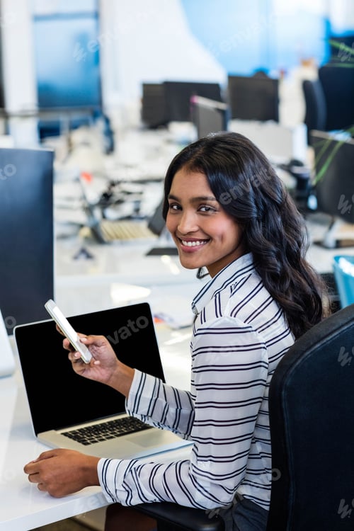Preview: Smartphone is resting beside laptop on white desk in modern open-plan office with monitors, cables