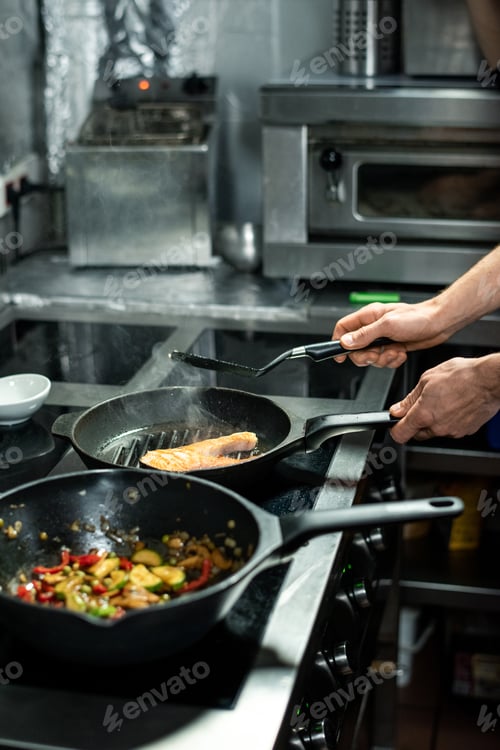 Preview: Hands of male chef roasting piece of salmon while cooking fish and vegetables