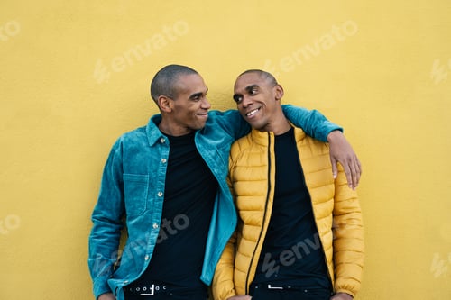Preview: African American adult identical twin brothers hugging and smiling, standing over yellow wall