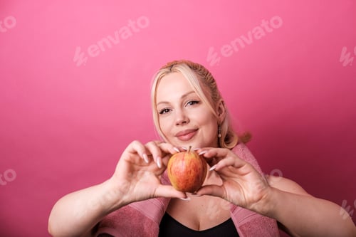 Preview: Sportswoman holding an apple promoting healthy lifestyle on pink background