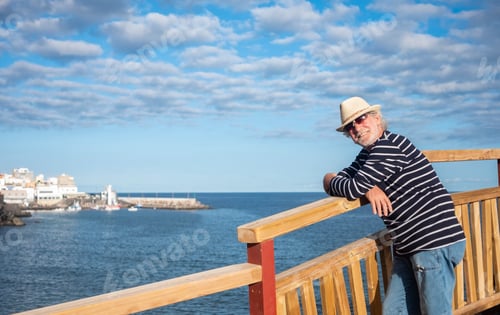 Preview: Smiling senior man with straw hat and blue striped shirt in sea excursion, harbour and blue sky