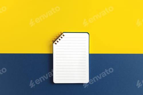 Preview: Top view of open notepad with white ruled paper page on yellow and blue background.