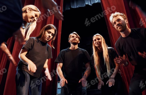 Preview: Standing against red curtains. Group of actors in dark colored clothes on rehearsal in the theater