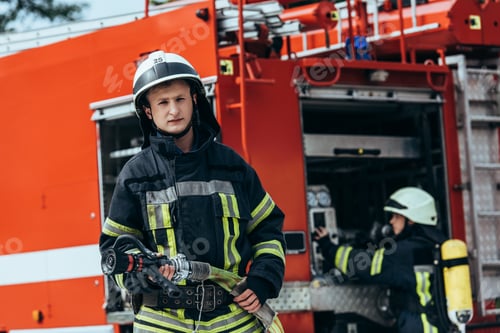 Preview: male firefighter in uniform holding water hose while colleague checking equipment in truck on street