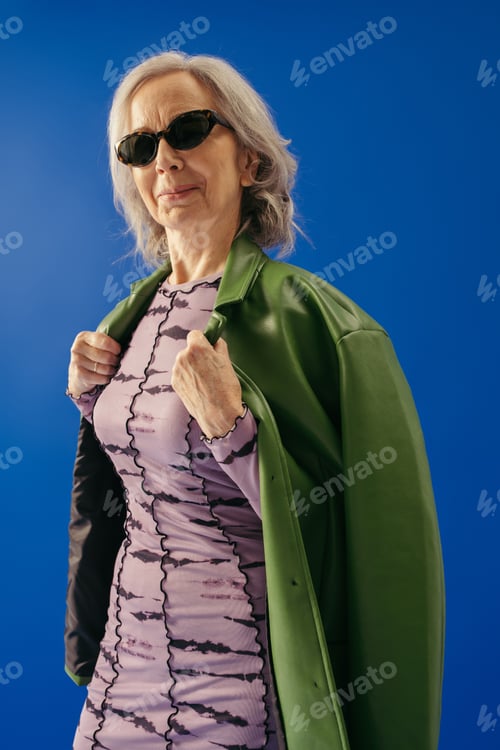 Preview: satisfied senior woman in sunglasses wearing green leather jacket isolated on blue
