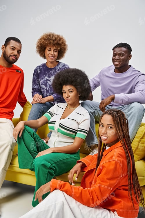 Preview: positive african american community in bright casual wear sitting together on yellow couch on grey