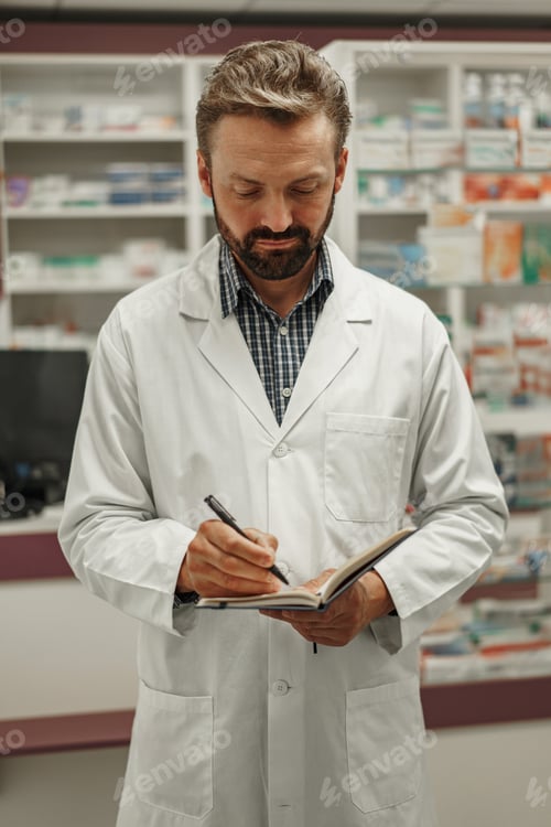 Preview: Focused male pharmacist in uniform is making notes in notepad while work in pharmacy