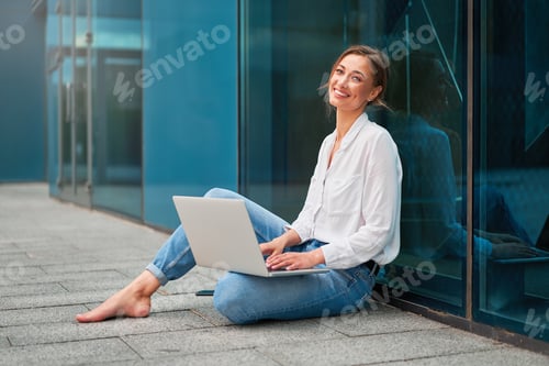 Preview: Blonde business woman is sitting on the floor barefoot with a laptop on her lap, smiling and looking