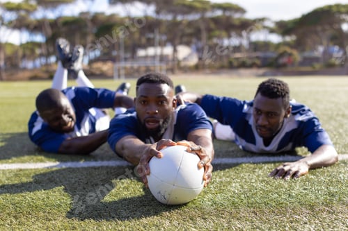 Preview: Three African American young male athletes reaching for a rugby ball on a grass field