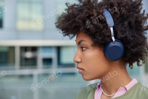 Preview: Sideways shot of curly haired Afro American woman listens music in headphones enjoys interesting plo