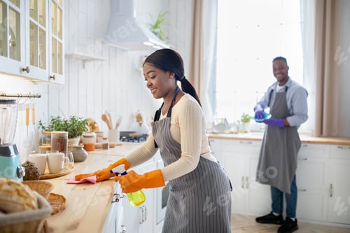 Preview: Cheerful black woman cleaning kitchen table, her boyfriend washing dishes on background, blank space
