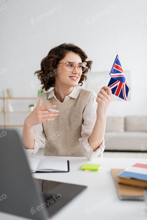 Preview: cheerful language teacher looking at flag of United Kingdom while sitting near blurred laptop at
