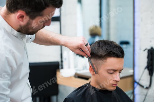 Preview: Portrait of handsome man with black hair having haircut at salon. Hairdresser holding comb and