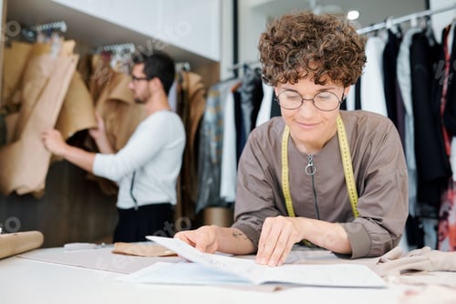 Preview: Young female seamstress or fashion designer looking at sketch of new model