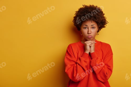 Preview: Photo of disappointed curly young woman keeps hands under chin, looks sadly at camera, feels lonelin