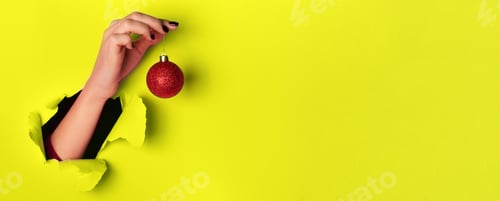 Preview: Woman holding red glittering Christmas ball in hand over yellow background with snow, light bokeh