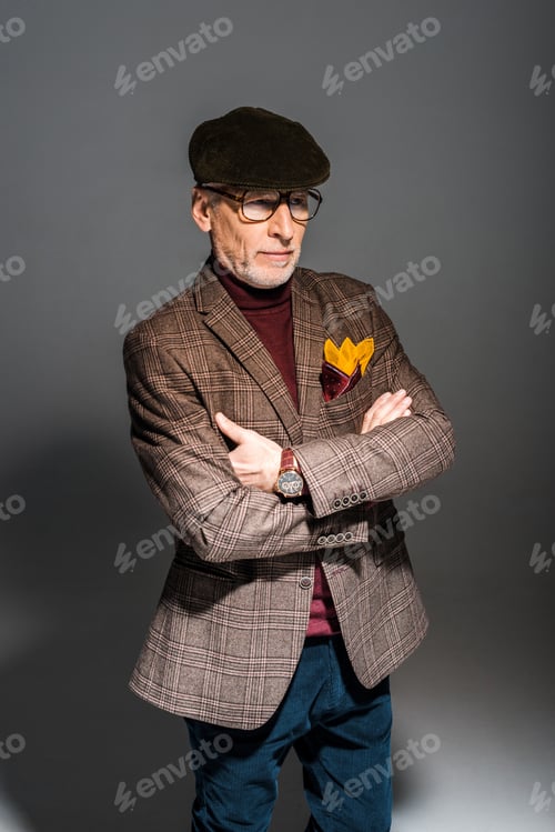 Preview: trendy middle aged man in glasses and cap standing with crossed arms on grey