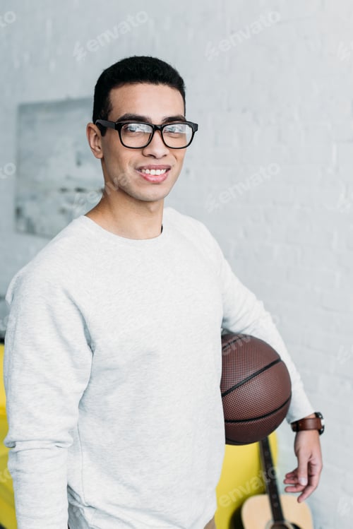 Preview: young mixed race man in white sweatshirt and glasses holding ball and looking at camera