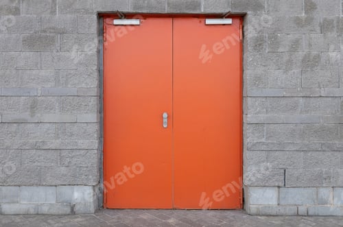 Preview: Gray brick wall with wide bright orange door in the centre of the photo. Entry into building