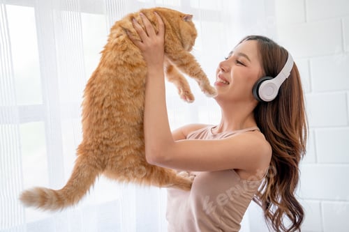 Preview: Pretty Asian girl wear earphone and hold cat in front of glass windows with white curtain