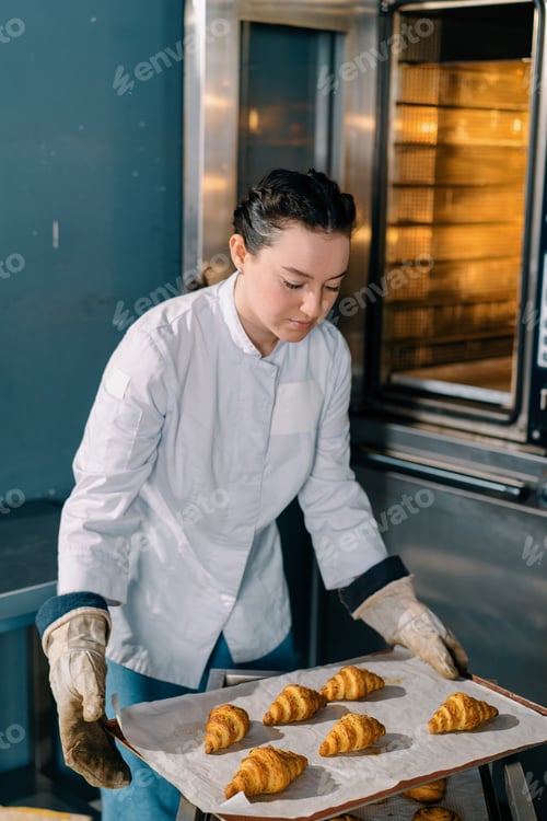 Preview: attractive baker takes ready buns and croissants from professional oven bakery production