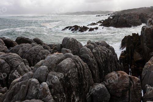 Preview: Jagged rocks and the rocky coastline of the Atlantic at De Kelders beach, waves breaking on shore.