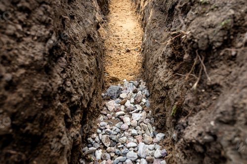 Preview: A trench dug in the yard along the fence to lay the drainage pipe,visible gravel and yellow sand.