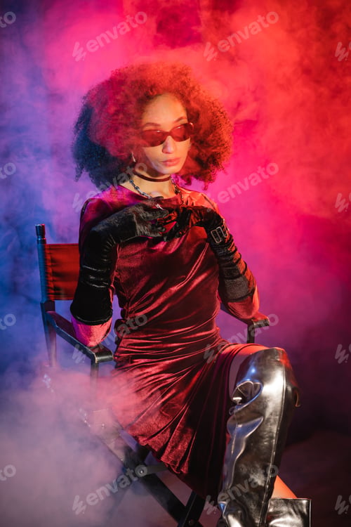 Preview: Trendy african american woman in dress and sunglasses sitting on chair on blue and red background