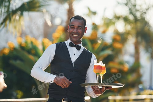 Preview: Holds cocktails. Black waiter in formal clothes is at his work outdoors at sunny daytime