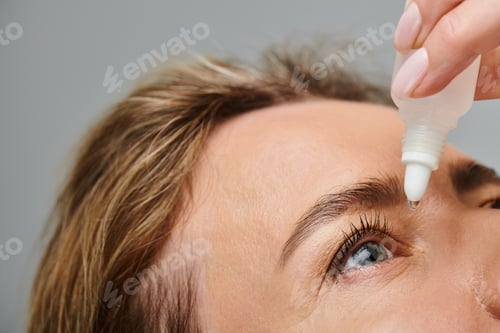 Preview: close up of attractive female model with blonde hair putting in eye drops while on gray backdrop