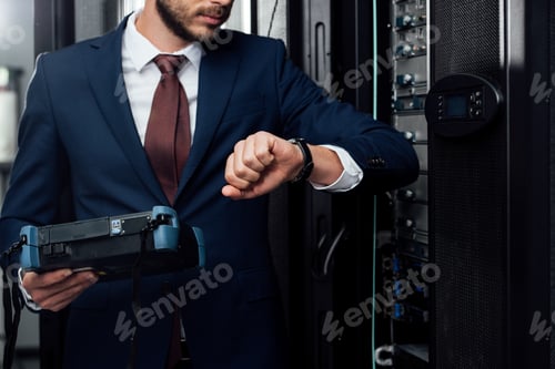 Preview: cropped view of businessman standing with reflectometer and looking at watch in server room