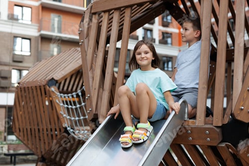 Preview: Happy and cheerful children, a boy and a girl, roll down a slide on a modern playground.