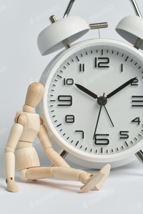 Preview: Wooden man sits near a white alarm clock. Concept of being late, long wait