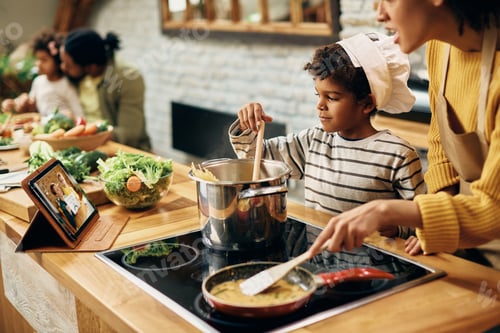 Preview: Smiling black boy cooking with is mother and following recipe on touchpad in the kitchen.