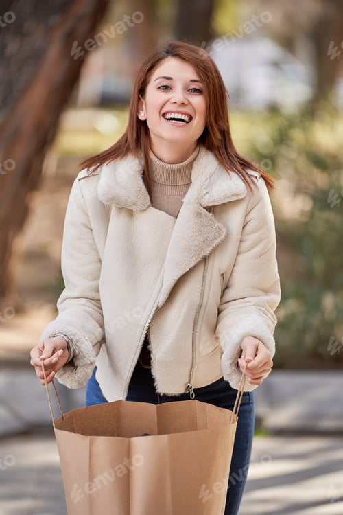 Preview: Woman looking at camera and smiling while holding a shopping bag outdoors.