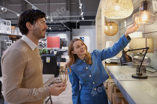 Preview: Young couple actively exploring various trendy lighting options in contemporary furniture store