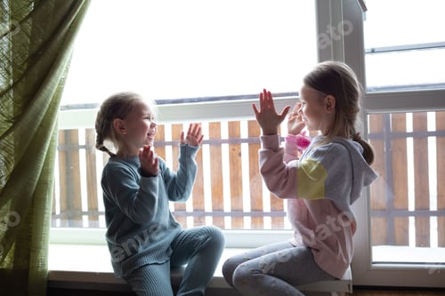 Preview: two little cute girls are playing patty-cake, sitting on the windowsill near the big window.