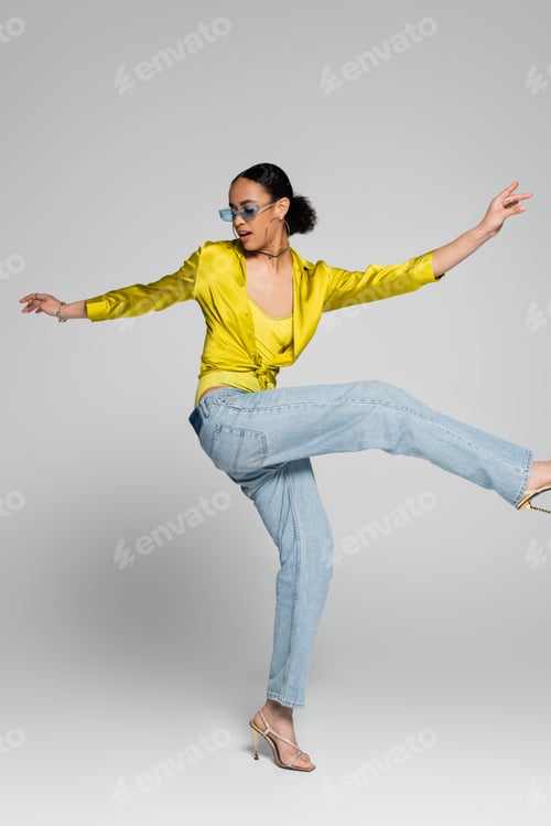 Preview: full length of excited african american woman in stylish outfit gesturing while posing on grey