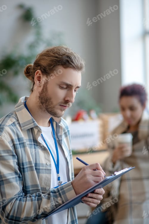 Preview: Young focused man with a badge standing and writing.