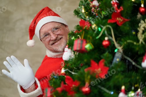 Preview: Senior man wearing Santa Claus costume surprise behind Christmas tree